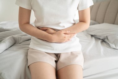Genç bir kadın evdeki yatakta otururken karın ağrısı çekiyor. Karın ağrısının nedenleri arasında adet sancısı, mide iltihabı, mide ülseri, gıda zehirlenmesi, ishal veya bağırsak iltihabı yer alıyor..
