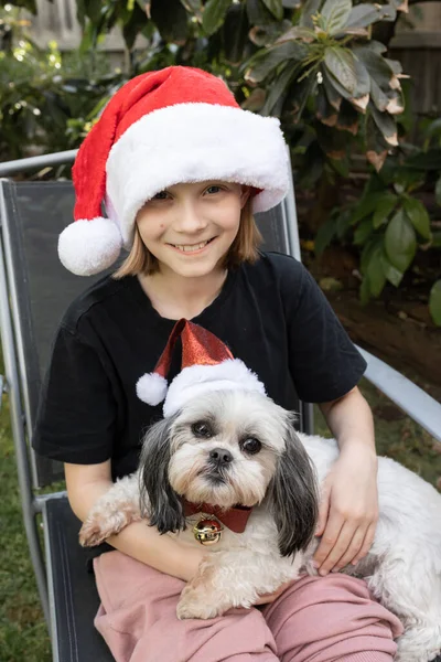A blonde caucasian australian girl wearing a Santa had while holding her pure bred grey and white Lhasa Apso dog which is wearing a Christmas hat and a red bow collar sitting on a chair outside