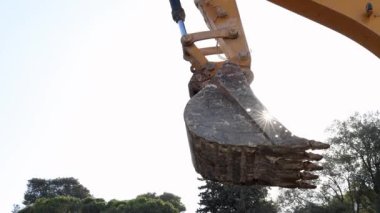 Manzaradaki ağaçlar ve suyla çevrili bir inşaat sahasında çalışan bir kazıcı kovasının detaylı bir yakın çekimi.