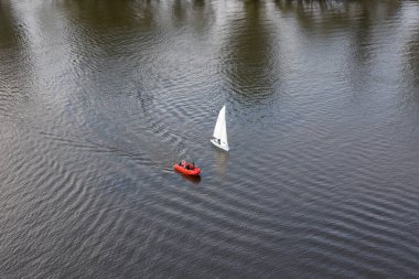 Küçük beyaz yelkenli ve kırmızı motorlu tekne geniş ve dalgalı bir nehirde yol alırlar. Yansımalar karanlık su yüzeyini bozar..