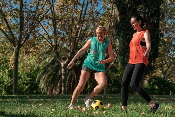 Two 30s years old woman training to play soccer or European football in amateur team. Yellow ball, sport field with green grass.
