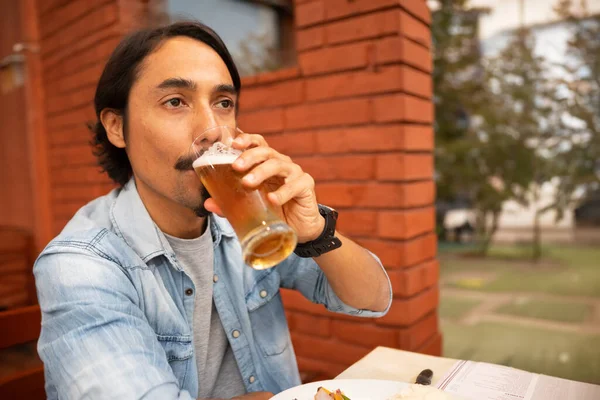 a latin man drinks beer sitting at a table on the terrace of his house.