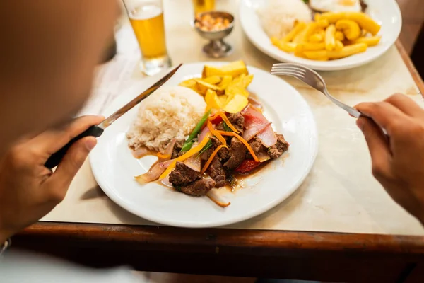 An unrecognizable person about to eat a lomo saltado, a typical Peruvian dish. Tenderloin steak cut in pieces, french fries, sarsa and rice. accompanied by cancha serrana, beer