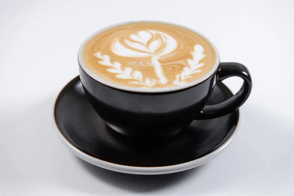 Süt köpüğünden çiçekli sıcak latte içeceği porselen bardakta ve kafede çay tabağıyla.