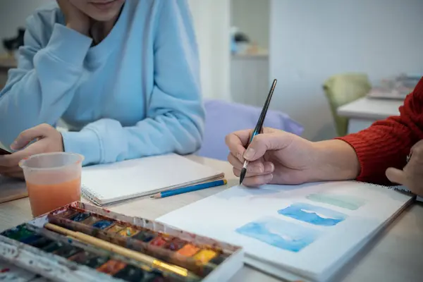 Sanat okulunda öğretmeniyle resim dersi alan bir çocuğun görüntüsü. Boyacı el fırçasıyla boya ve masada resim çizer..