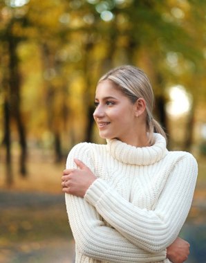 Sonbahar parkında beyaz kazaklı güzel sarışın bir kadın yürüyor. Parkta sonbahar yürüyüşleri. Sonbaharda aktif tatiller.