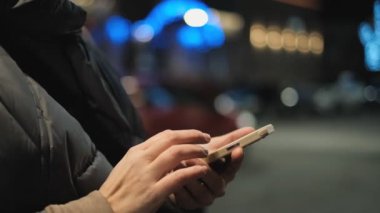 Bir kadın kışın akşamları sokakta duruyor ve arka planda Noel çelenkleri için akıllı telefon kullanıyor. Noel 'de akıllı telefon kullanarak iletişim.