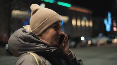 Bir kadın kışın akşamları sokakta duruyor ve arka planda Noel çelenkleri için akıllı telefon kullanıyor. Noel 'de akıllı telefon kullanarak iletişim.