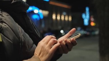 Bir kadın kışın akşamları sokakta duruyor ve arka planda Noel çelenkleri için akıllı telefon kullanıyor. Noel 'de akıllı telefon kullanarak iletişim.