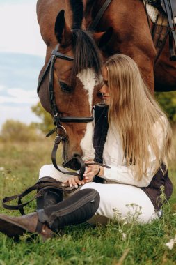 Young beautiful female jockey sits on a meadow near her horse at sunset. Walk with a horse in the summer on a meadow.