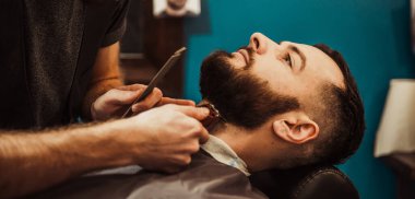 A professional barber cuts his beard to a young hipster man. Beard trim at the barbershop. Stylish beard cut.
