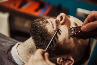 A professional barber cuts his beard to a young hipster man. Beard trim at the barbershop. Stylish beard cut.