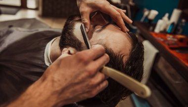 A professional barber cuts his beard to a young hipster man. Beard trim at the barbershop. Stylish beard cut.