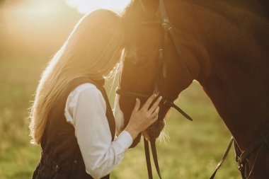 Young beautiful female jockey strokes and hugs the horse's head and prepares for the competition. Jumping training in the meadow in summer evening.
