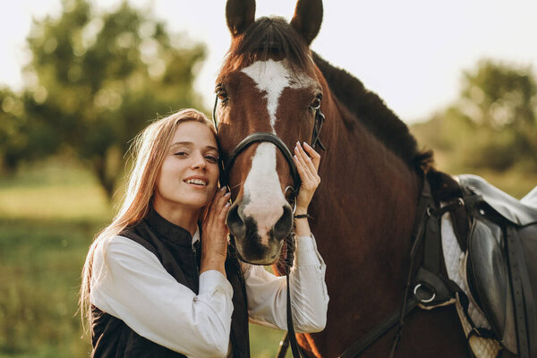 Young beautiful female jockey strokes and hugs the horse's head and prepares for the competition. Jumping training in the meadow in summer evening.