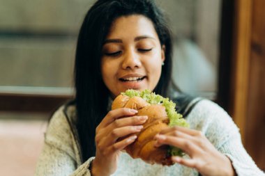 A young African American woman sits in a cafe and eats an appetizing burger. Bad cheap food. Wrong nutrition.
