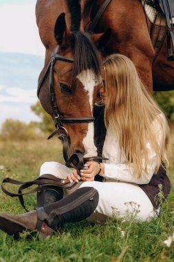 Young beautiful female jockey sits on a meadow near her horse at sunset. Walk with a horse in the summer on a meadow.