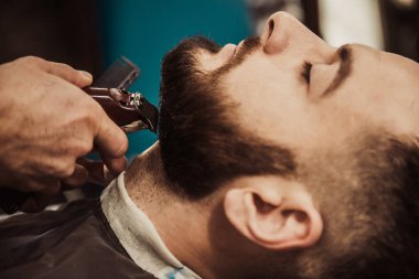 A professional barber cuts his beard to a young hipster man. Beard trim at the barbershop. Stylish beard cut.