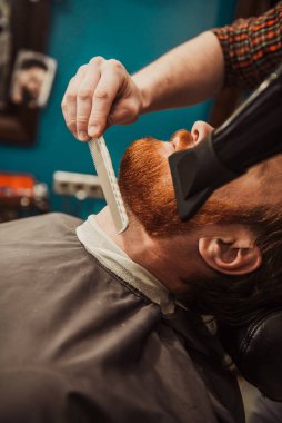 A professional barber cuts his beard to a young hipster man. Beard trim at the barbershop. Stylish beard cut.