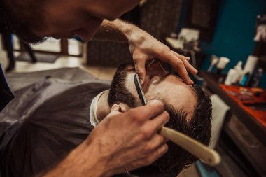 A professional barber cuts his beard to a young hipster man. Beard trim at the barbershop. Stylish beard cut.