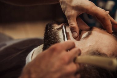 A professional barber cuts his beard to a young hipster man. Beard trim at the barbershop. Stylish beard cut.