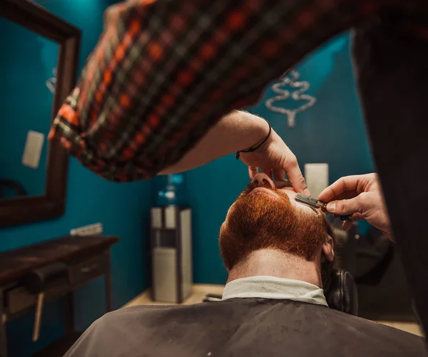 A professional barber cuts his beard to a young hipster man. Beard trim at the barbershop. Stylish beard cut.