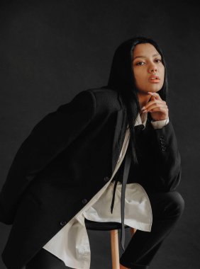 Genç bir Afrikalı Amerikalı kadın siyah bir arka planda iş elbisesi içinde bir sandalyede oturuyor. Gösterişçi genç bir zenci kadın..