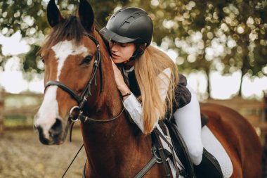 A young woman jockey lies on her horse and hugs her. Preparing for the show jumping championship.