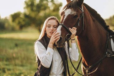 Young beautiful female jockey strokes and hugs the horse's head and prepares for the competition. Jumping training in the meadow in summer evening.