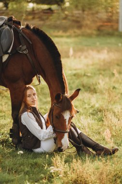 A young beautiful woman jockey with her dog sits in a meadow near her horse at sunset. Walk with a horse in the summer on a meadow.