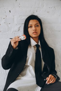 Koyu tenli genç bir kadın beyaz bir odada iddialı bir şekilde oturur ve yemek çubuklarıyla suşi yer. Takım elbiseli Afrikalı Amerikalı kadın rulo yiyor..