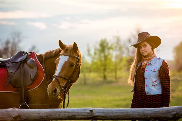 cowgirl in a hat standing near a horse in a field
