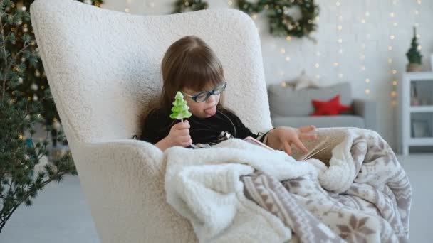 ダウン症の少女が椅子に座って子供の本を見て クリスマスイブで笑いを食べる — ストック動画
