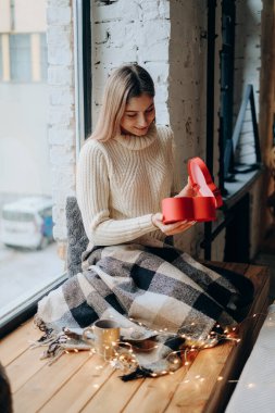 Young beautiful blonde opens a gift for Valentine's Day. A woman opens a red box in the form of a heart with a gift.