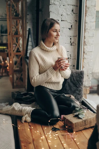 Genç ve güzel bir kadın bulutlu bir sabahta elinde bir fincan kahveyle pencere eşiğinde oturuyor. Bulutlu bir sabahta sıcak kahve.