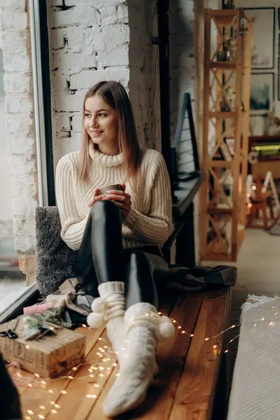 Genç ve güzel bir kadın bulutlu bir sabahta elinde bir fincan kahveyle pencere eşiğinde oturuyor. Bulutlu bir sabahta sıcak kahve.