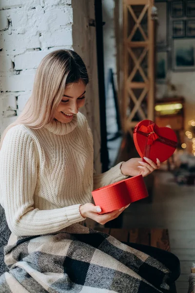 Young beautiful blonde opens a gift for Valentine's Day. A woman opens a red box in the form of a heart with a gift.
