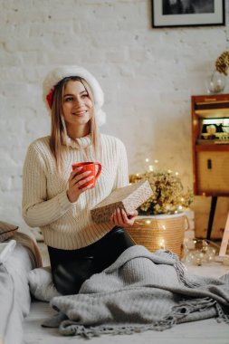 Genç ve güzel bir kadın, Noel arifesinde pencerenin yanındaki bir evde elinde bir kupa sıcak çayla oturuyor..