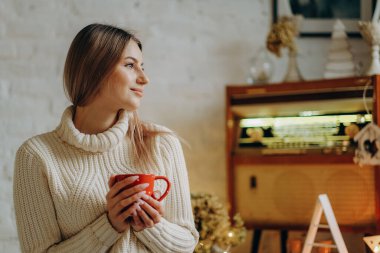 Genç ve güzel bir kadın, Noel arifesinde pencerenin yanındaki bir evde elinde bir kupa sıcak çayla oturuyor..