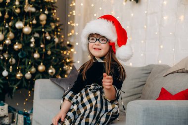 Down sendromlu küçük bir kız kanepeye oturmuş Noel Baba şapkası takıyor ve Noel hediyelerini bekliyor. Down sendromlu bir çocuğu büyütmekte güçlük çekiyorlar..