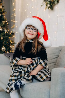 Down sendromlu küçük bir kız kanepeye oturmuş Noel Baba şapkası takıyor ve Noel hediyelerini bekliyor. Down sendromlu bir çocuğu büyütmekte güçlük çekiyorlar..