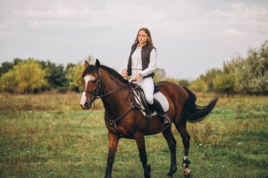 Young beautiful blonde woman jockey rides a brown horse in a meadow at sunset in summer. Preparing for an equestrian competition.