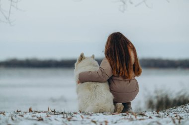 Kışın sevgili evcil hayvanı Samoyed ile parkta bir gölün kıyısında yürüyen bir kız. Kışın köpeği gezdiriyorum..