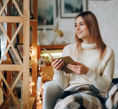 Genç, güzel bir kadın, battaniyeyle kaplı bir sandalyede oturur ve Noel arifesinde pencerenin yanında kitap okur. Noel arifesinde rahat bir sandalyede kitap okumak..
