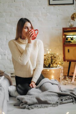 Genç ve güzel bir kadın, Noel arifesinde pencerenin yanındaki bir evde elinde bir kupa sıcak çayla oturuyor..