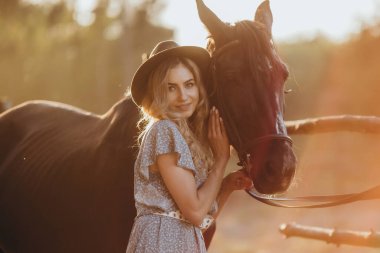 Atı olan genç bir kadın sonbaharda günbatımında çiftliğinde çitin yanında duruyor. Binicilik sporu için safkan atlar yetiştiriyor..