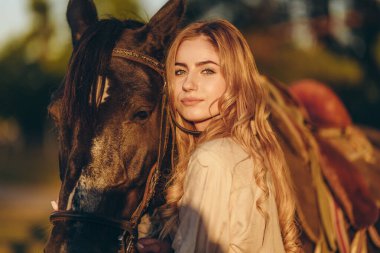 Atı olan genç bir kadın sonbaharda günbatımında çiftliğinde çitin yanında duruyor. Binicilik sporu için safkan atlar yetiştiriyor..