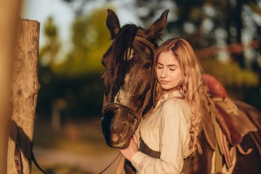 Atı olan genç bir kadın sonbaharda günbatımında çiftliğinde çitin yanında duruyor. Binicilik sporu için safkan atlar yetiştiriyor..
