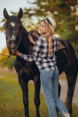 Atı olan genç bir kadın sonbaharda günbatımında çiftliğinde çitin yanında duruyor. Binicilik sporu için safkan atlar yetiştiriyor..