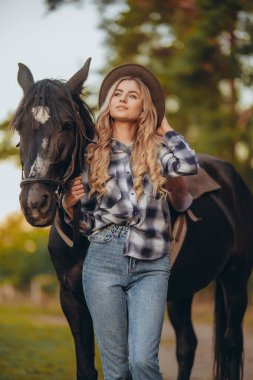 Atı olan genç bir kadın sonbaharda günbatımında çiftliğinde çitin yanında duruyor. Binicilik sporu için safkan atlar yetiştiriyor..